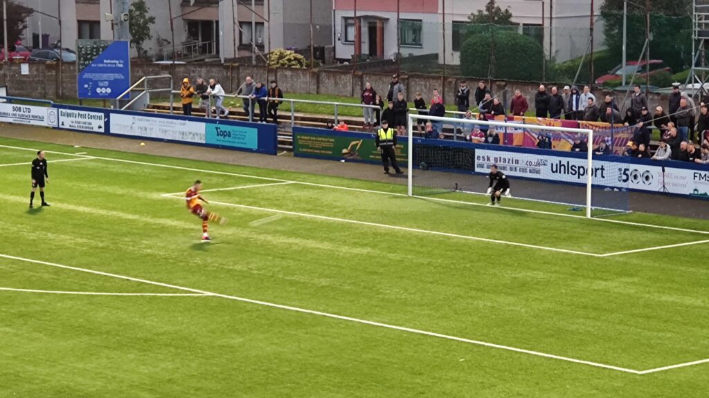 A Motherwell player scores during the shoot-out in front of the away end.